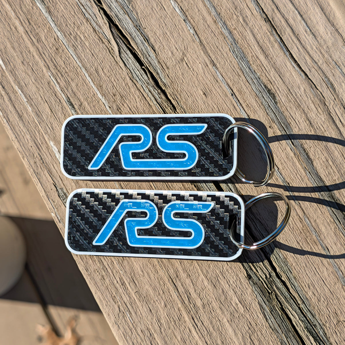 Two rectangular keychains with a carbon fiber pattern background and a blue 'RS' logo, resting on a wooden deck.