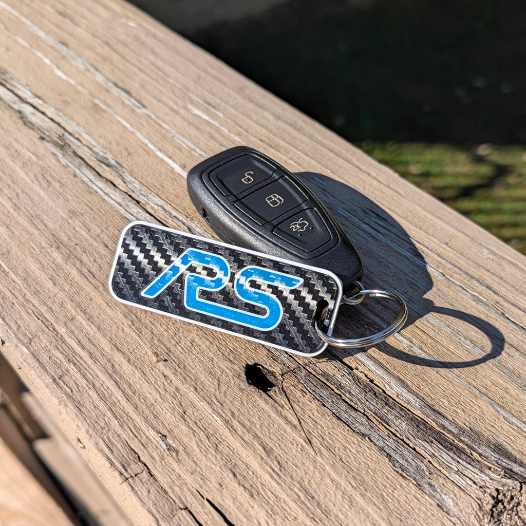 A carbon fiber patterned keychain with a blue 'RS' logo and a black car key fob resting on a weathered wooden deck in bright sunlight.
